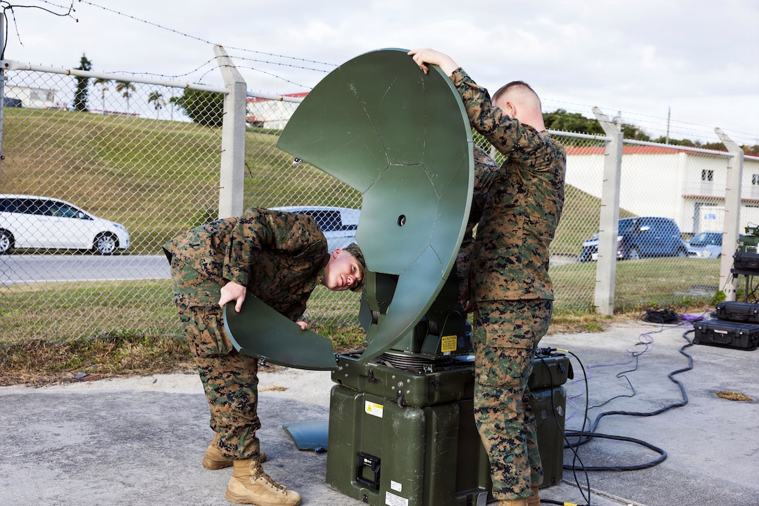 Two men wearing camouflage military uniforms assemble a large satellite on a paved surface next to a chain-link fence with a road and buildings in the background.