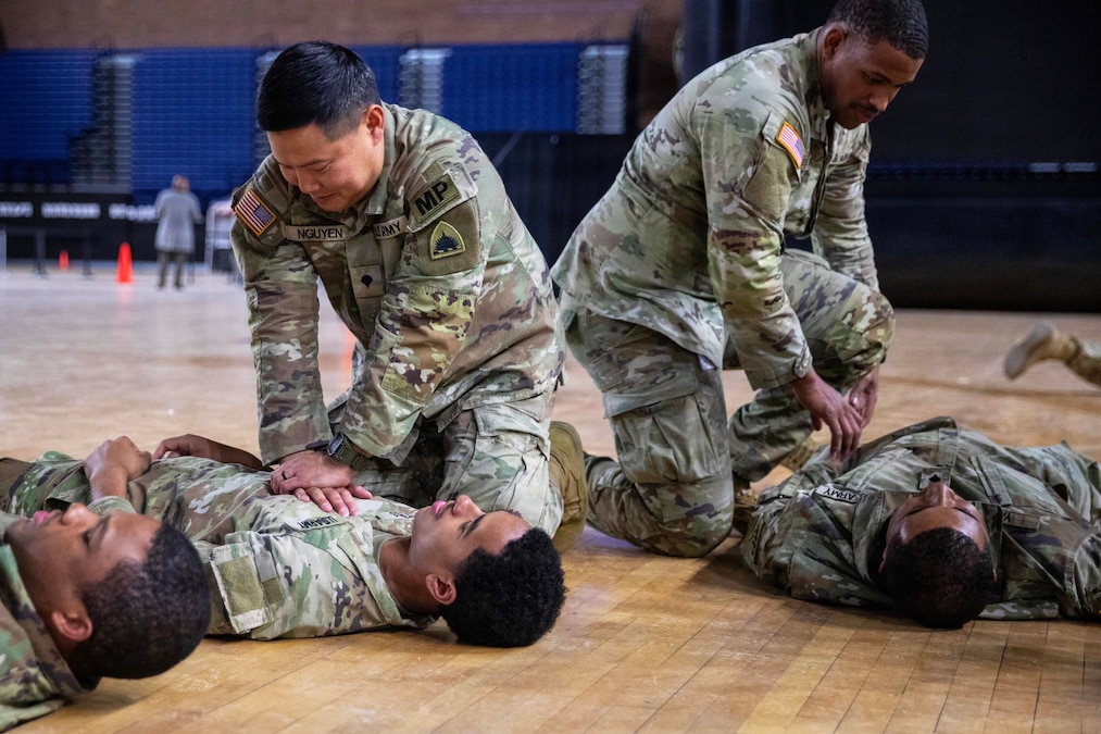 Three men wearing camouflage military uniforms lie on the floor of a gymnasium while two other men wearing similar attire perform CPR on two of the people lying down.