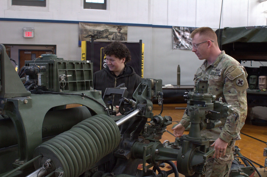 Sgt 1st Class James Nelson explains and assists AJ Spear in loading a dummy round into an M119A3 Howitzer at the Sycamore Armory.
