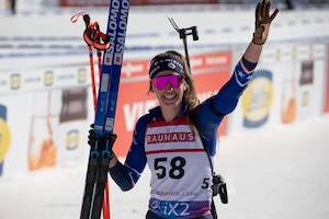 A woman wearing goggles and ski sports gear holds skis while smiling and waving.