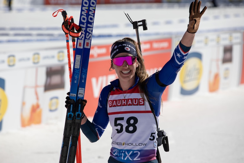 A woman wearing goggles and ski sports gear holds skis while smiling and waving.