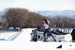A woman wearing goggles and ski sports gear holds ski poles while skiing across a snowy slope outdoors, with a crowd observing and snow-covered mountains in the background.