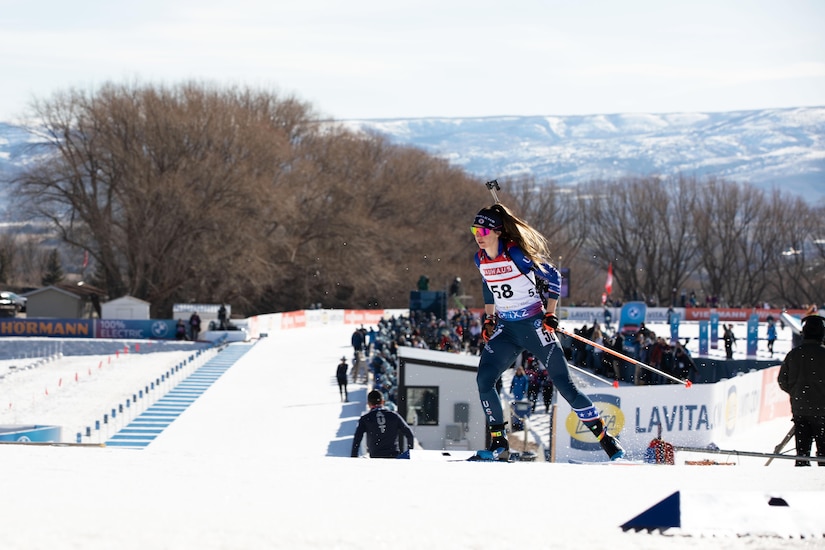 A woman wearing goggles and ski sports gear holds ski poles while skiing across a snowy slope outdoors, with a crowd observing and snow-covered mountains in the background.