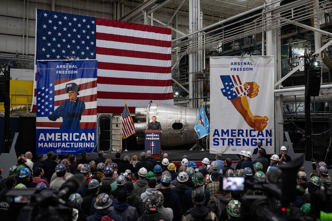 A standing crowd, many in hard hats, faces Secretary of War Pete Hegseth, who speaks from a lectern flanked by posters.
