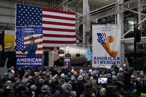A standing crowd, many in hard hats, faces Secretary of War Pete Hegseth, who speaks from a lectern flanked by posters.