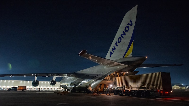 An aircraft sits on a flight line with its rear open at night, while about five people prepare to load a large storage container into the back.