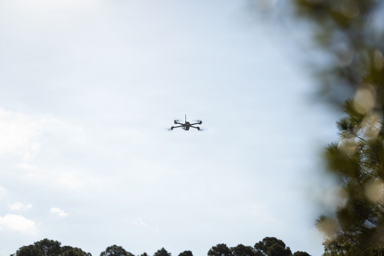 A drone flies in the air with blurry trees in the foreground.