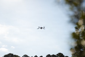 A drone flies in the air with blurry trees in the foreground.