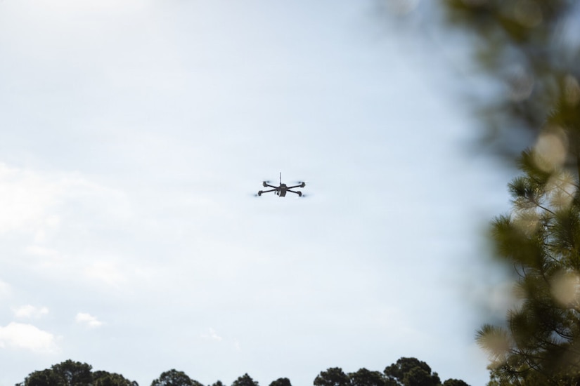 A drone flies in the air with blurry trees in the foreground.