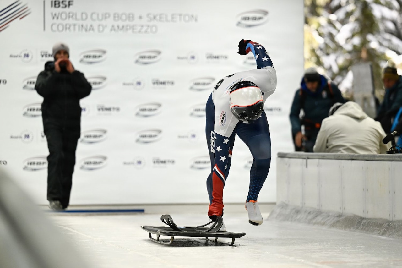 A woman dressed in winter Olympic sledding gear bends down toward a sled while on ice.