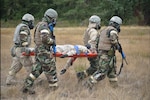 Four men in military uniform carry a patient in a field.