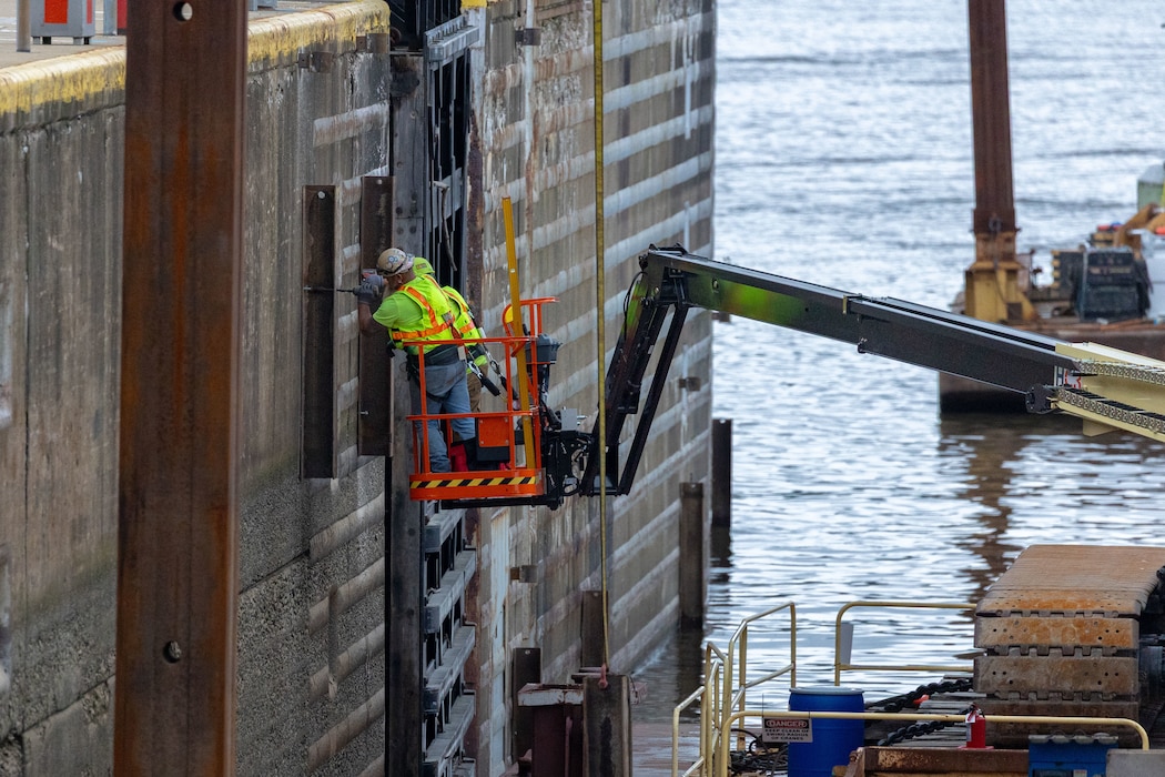 Two men in a crane use construction equipment to attach a beam to a river lock wall.