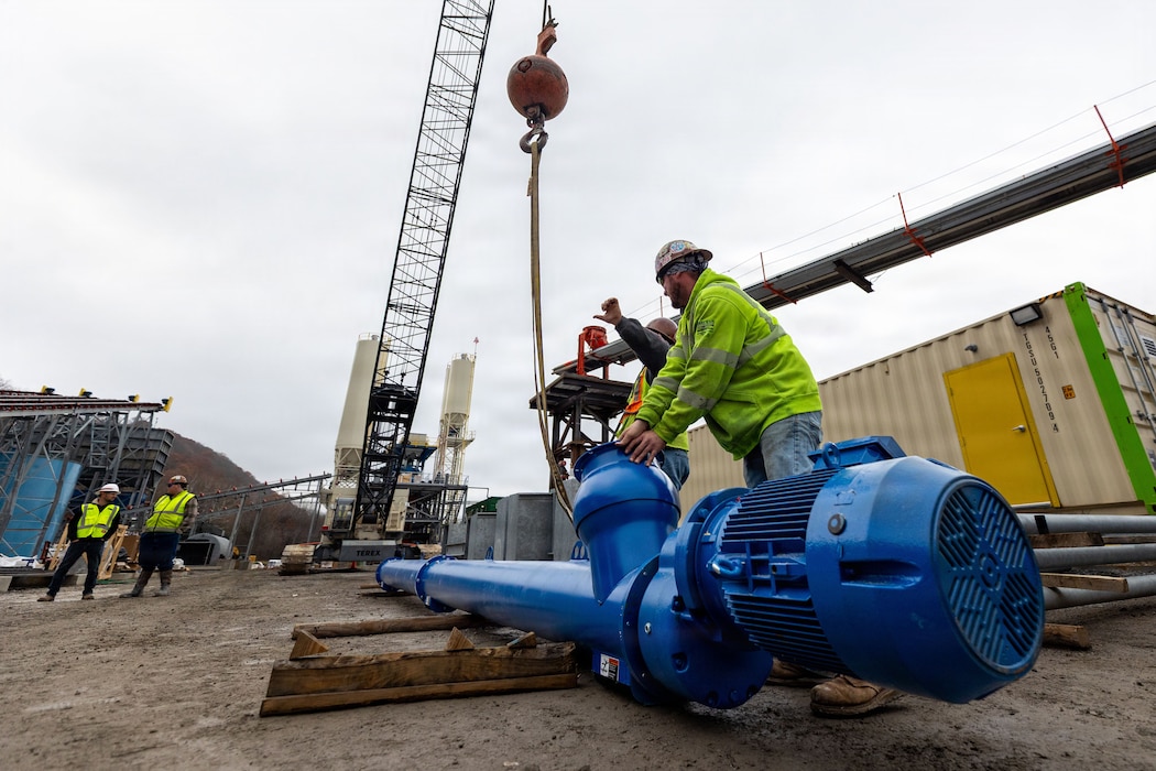 Two men at a construction site attach a cylindrical steel component to a crane.