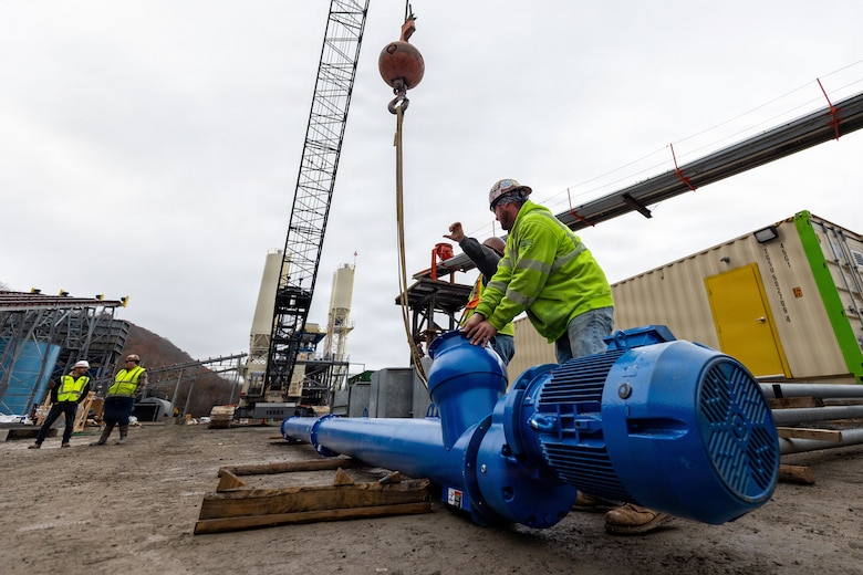 Two men at a construction site attach a cylindrical steel component to a crane.