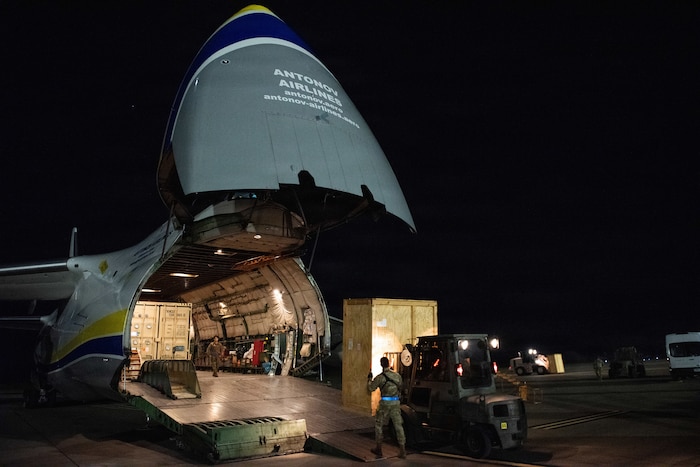 People guide a forklift carrying a wooden box into the open nose of a large aircraft at night.