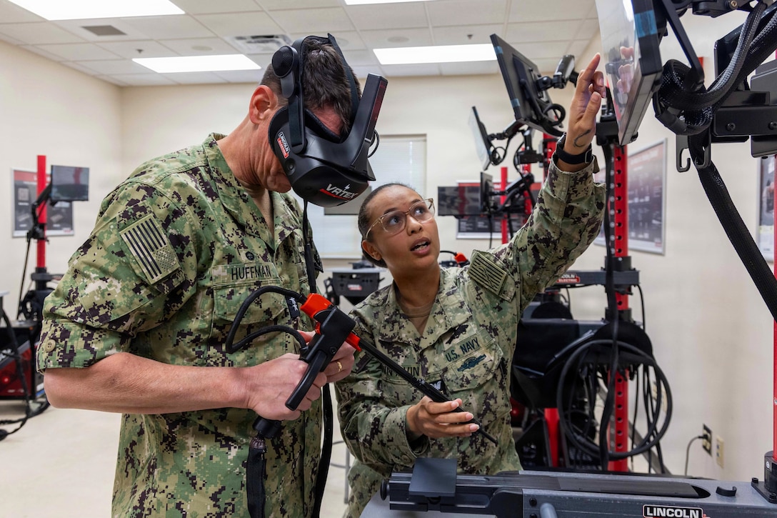 A man and a woman wearing camouflage military uniforms stand in a room with monitors and virtual reality equipment. The man is wearing a virtual reality headset and holding a virtual reality welding tool as the woman looks upward and points at a monitor.
