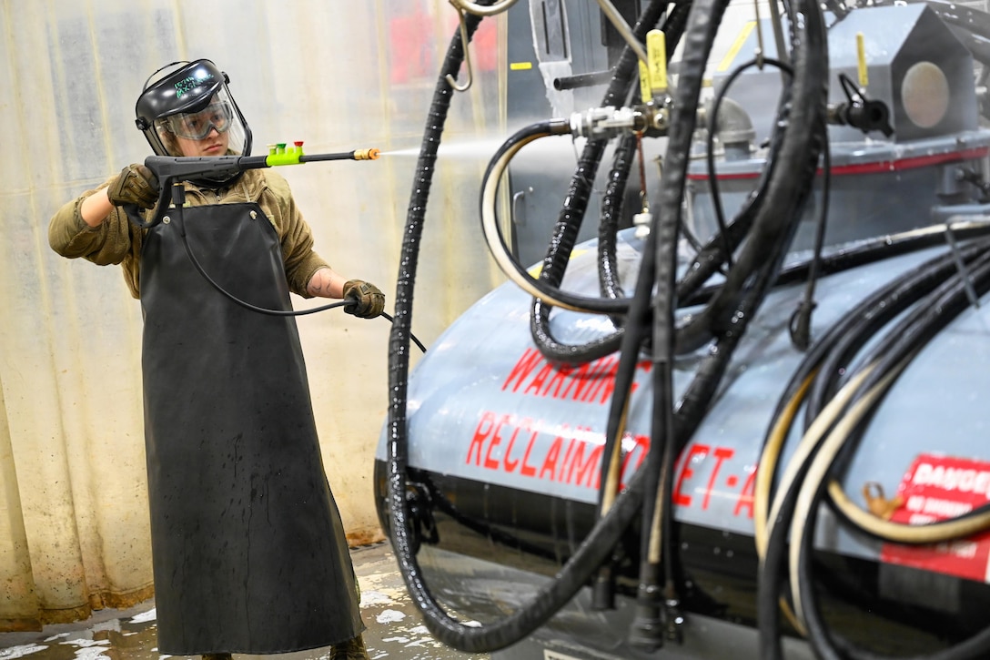 A person wearing a face shield, gloves and a long waterproof apron uses a water spray gun to clean equipment; numerous hoses are in the foreground.