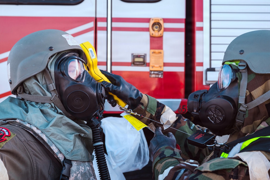 A person wearing a helmet and respirator holds a digital reader on the head of another person in similar attire.