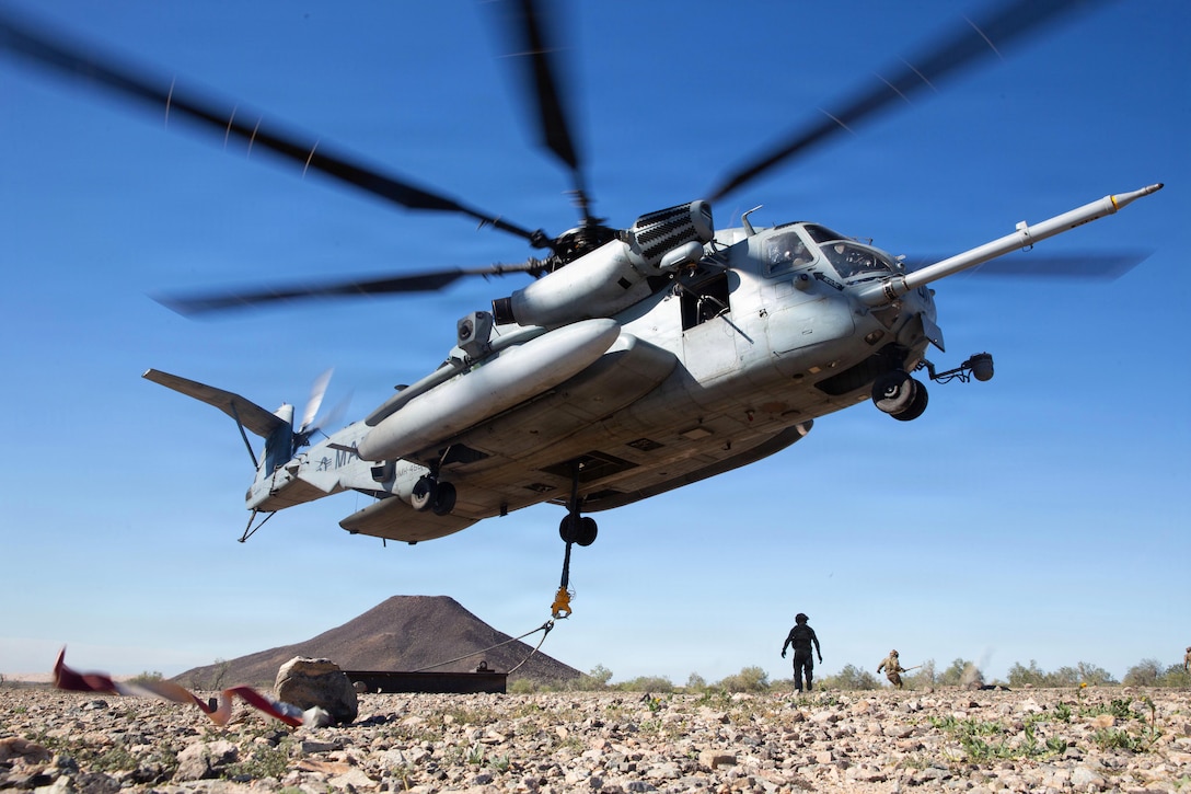 A helicopter hovers in a blue sky over a rocky area with a mountain in the background while being hooked to a flatbed; two people observe in the distance.