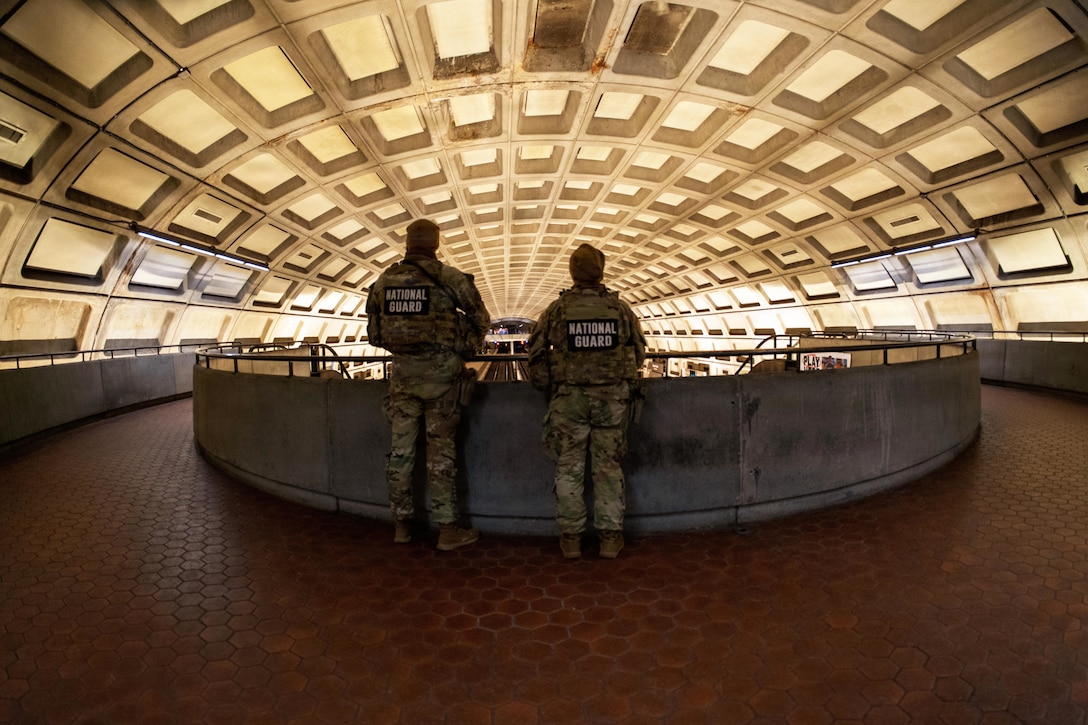 Two people wearing camouflage military uniforms and covers stand side by side inside a train station, observing the people below from an elevated walkway.