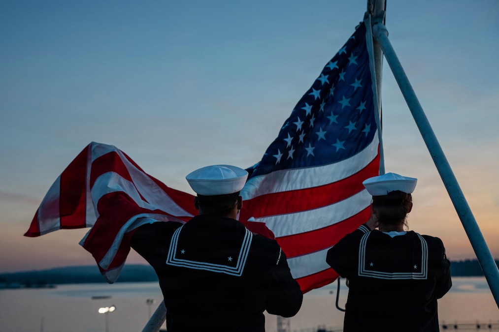Two people wearing dark blue military uniforms and white hats face backward while holding an American flag attached to a pole outside under a pastel sky.
