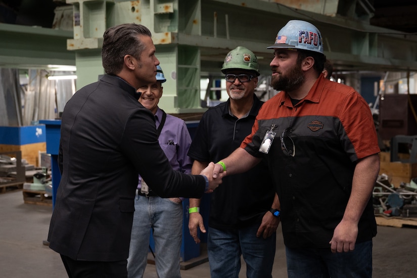 Secretary of War Pete Hegseth shakes hands with a person wearing a hard hat as others look on in an industrial-type space.