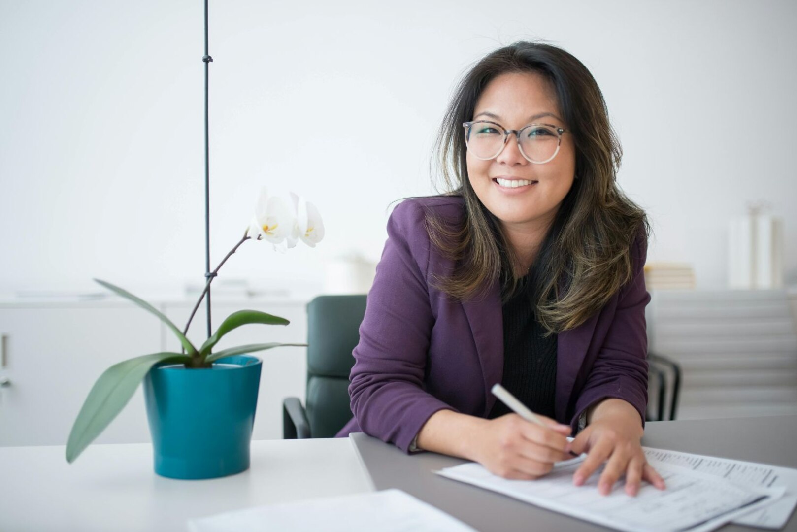 Smiling woman wearing glasses sits at table with potted plant.