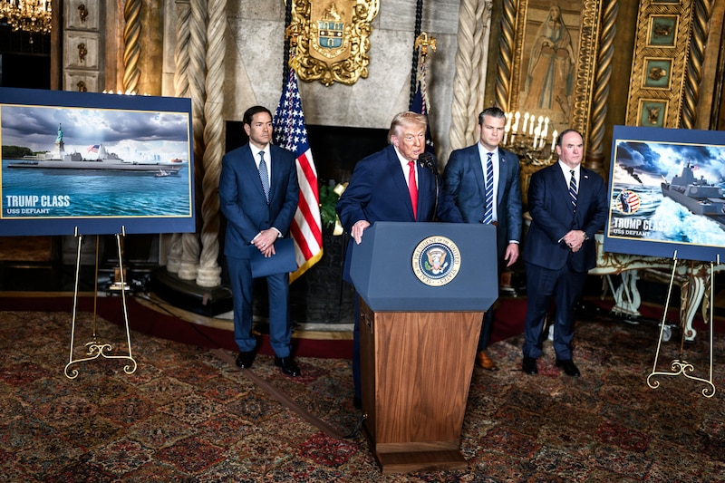 A man in a suit with a red tie talks from a lectern indoors as another man stands behind him.