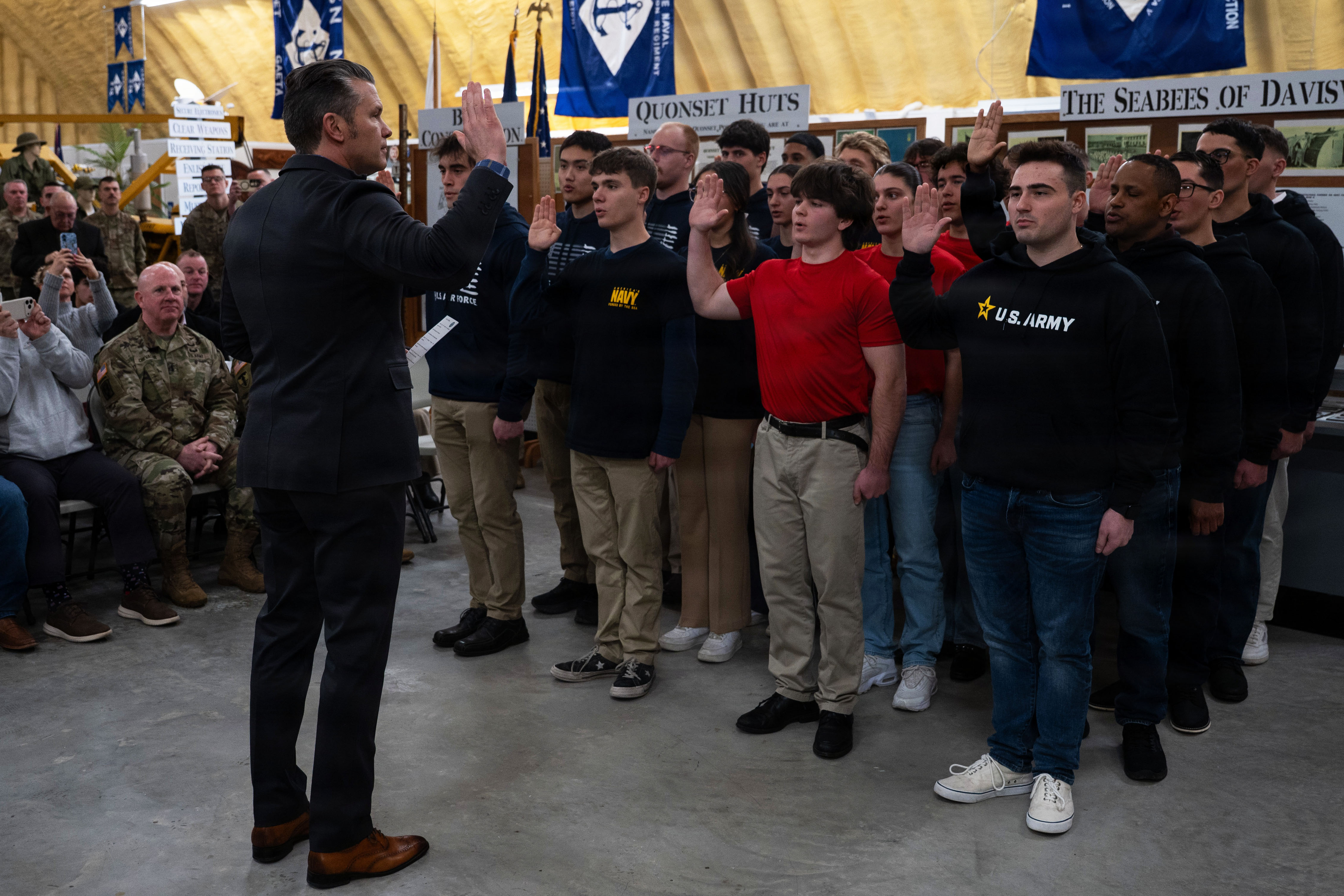 A man in business attire raises his right hand and addresses people standing in front of him, also with their right hands raised, as other people watch from the side, in a large room.