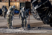People in protective gear look at gravel on the ground while holding shovels.