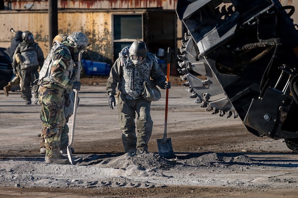 People in protective gear look at gravel on the ground while holding shovels.