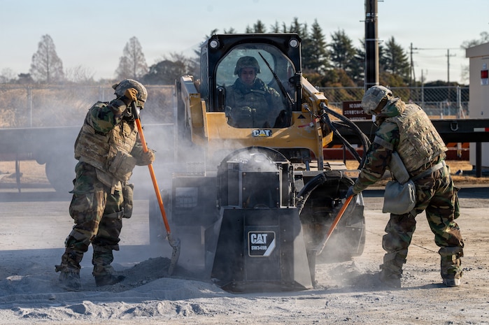 Airmen dressed in personal protective chemical gear operate construction equipment and use shovels.