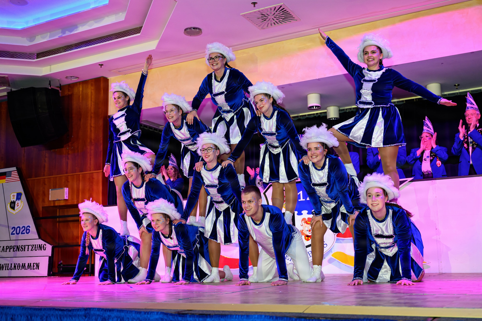 A team of dancers form a human pyramid on a stage inside a ballroom.