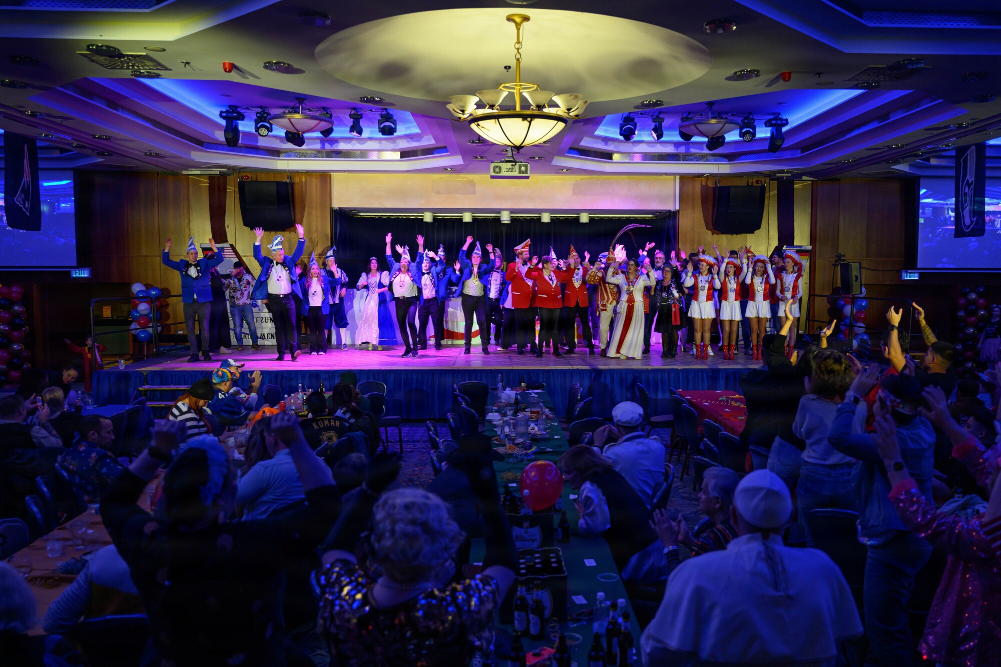 A group of people dressed in costumes dance on a stage inside a ballroom.