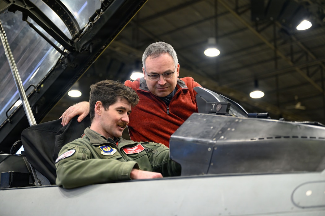 A pilot sitting in a cockpit speaks to a man standing next to him on a ladder.