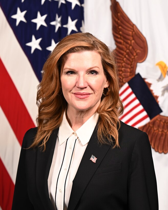 White woman with strawberry blonde hair poses for a photo. She is wearing a white collared button up shirt with a black blazer over with a U.S. flag pin pinned on her left blazer lapel.
She is posed looking directly at the camera with the United States behind her.