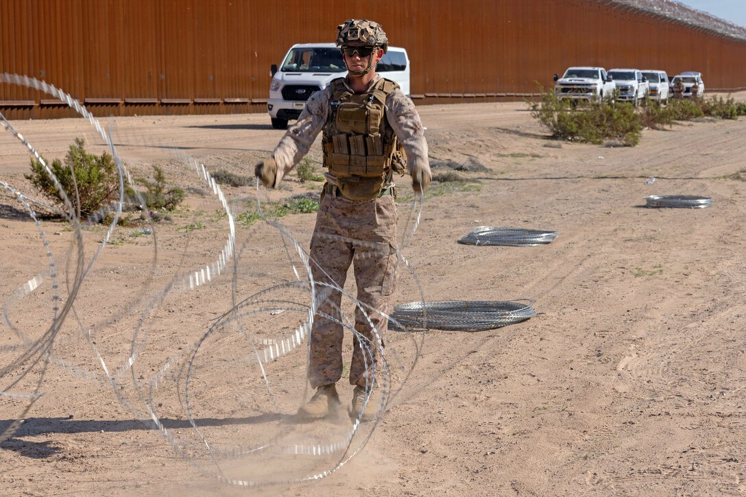 A Marine uncoils concertina wire on flat arid landscape near a tall wall, with wire coils on the ground nearby.