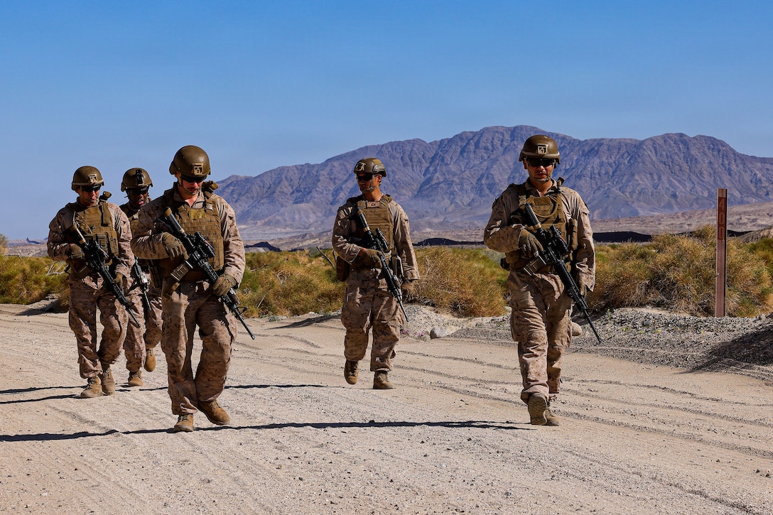 Five Marines carrying weapons walk on a sandy roadway in arid landscape with mountains in the background.
