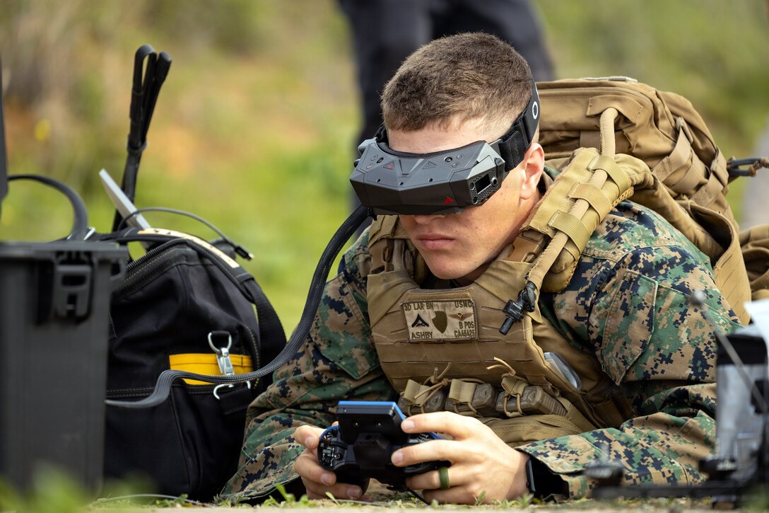 A Marine operates a hand-held device next to a piece of equipment  in a wooded area while wearing black eye gear.