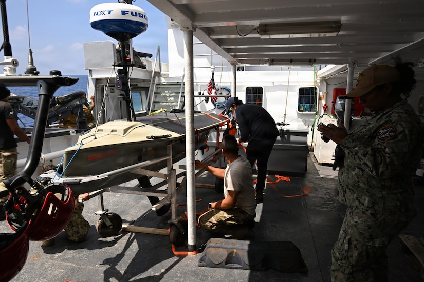 Two men wearing casual attire work on an unmanned surface vessel aboard a large ship. A woman in a camouflage military uniform on the right is looking at a device in her hands. There are four other people wearing camouflage military uniforms and casual attire working around the unmanned vessel in the background.