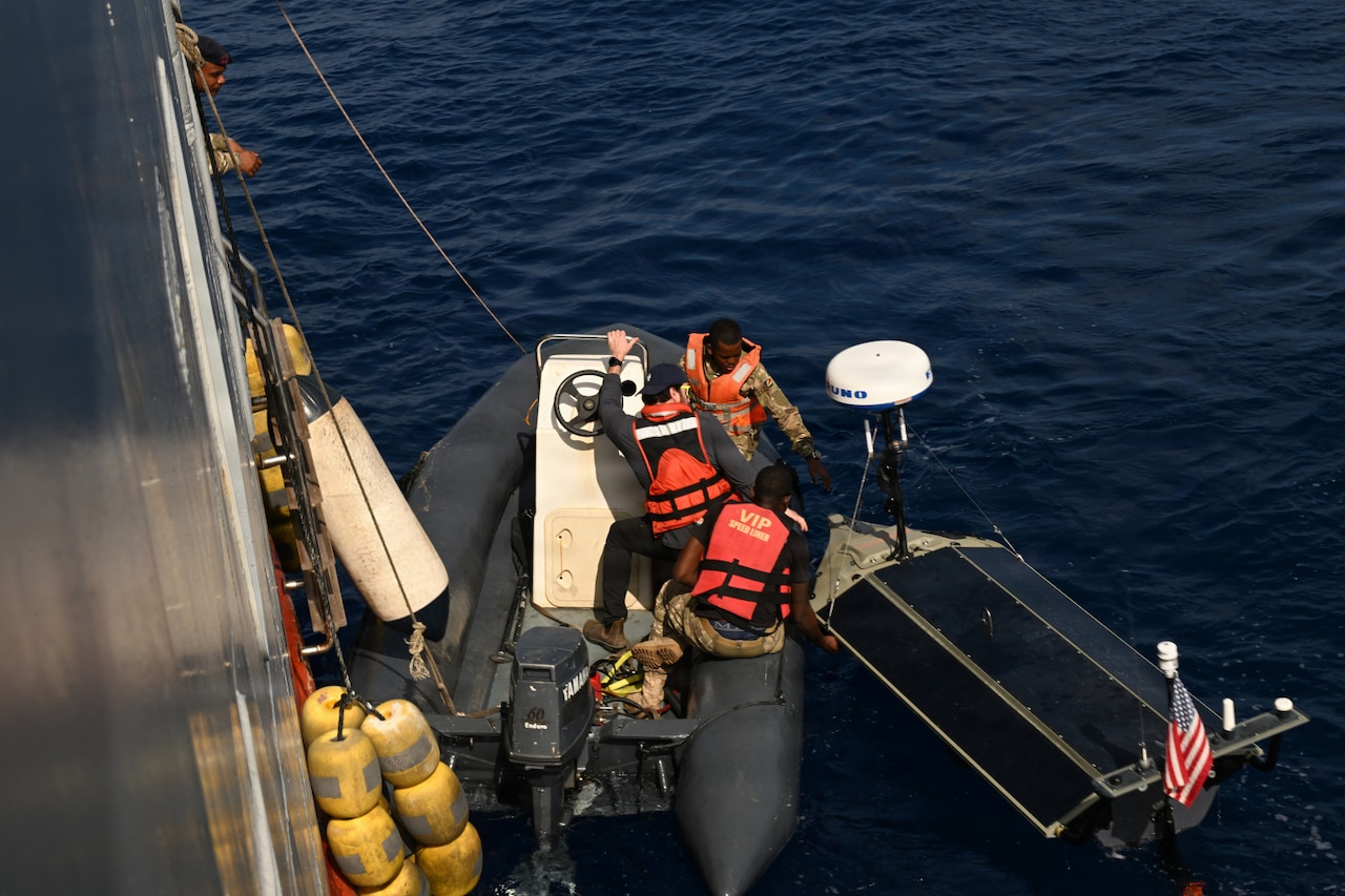 Two men wearing camouflage military uniforms and life jackets and another man in casual attire and a life jacket launch an unmanned surface vessel from a small boat in the water. The boat has ropes attached to it from a large ship on the left.
