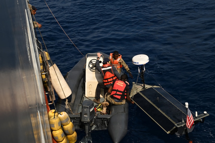 Two men wearing camouflage military uniforms and life jackets and another man in casual attire and a life jacket launch an unmanned surface vessel from a small boat in the water. The boat has ropes attached to it from a large ship on the left.