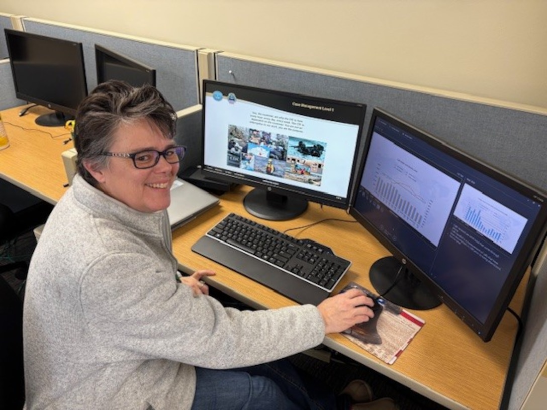 A woman with short, dark hair and glasses is seated at a desk in a cubicle, smiling at the camera. On the desk in front of her are two computer monitors displaying presentation slides with graphs and images.