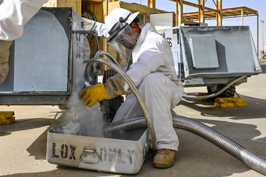 A man wearing a white coverall, a hard hat with a face shield attached and gloves kneels on rocky terrain near metal equipment as white vapor emits from it. More equipment and hoses are in the background.