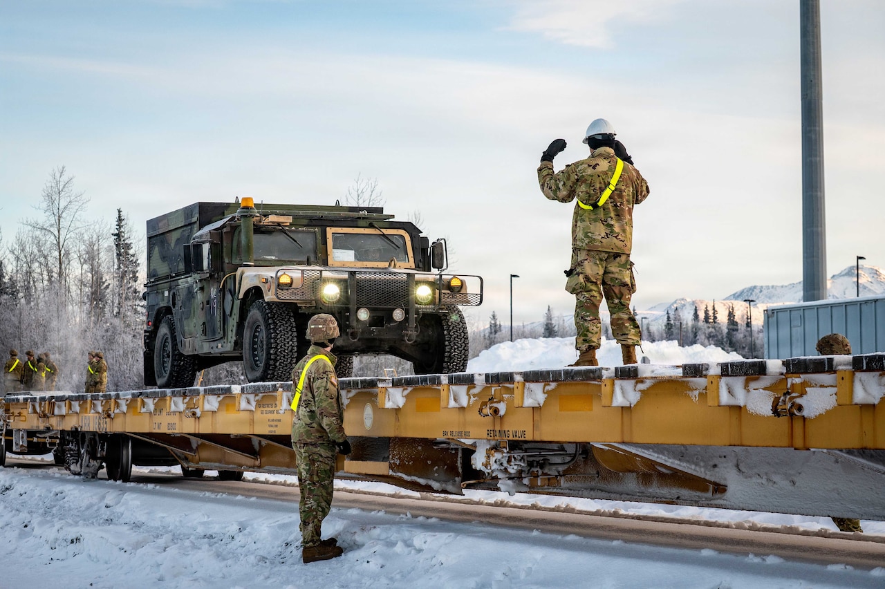 A person wearing a camouflage military uniform and a hard hat stands on a flatcar and gestures with his hands to guide a military vehicle; another person in similar attire stands on the ground and observes.