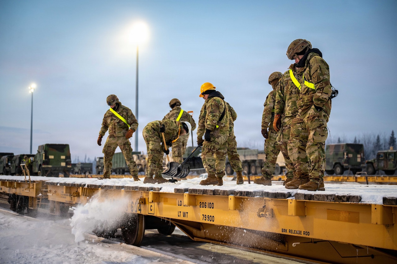 About eight people wearing camouflage military uniforms, safety belts and helmets remove snow from a flatcar at a railhead outside under a blue sky with tower lights and other military vehicles in the distance.