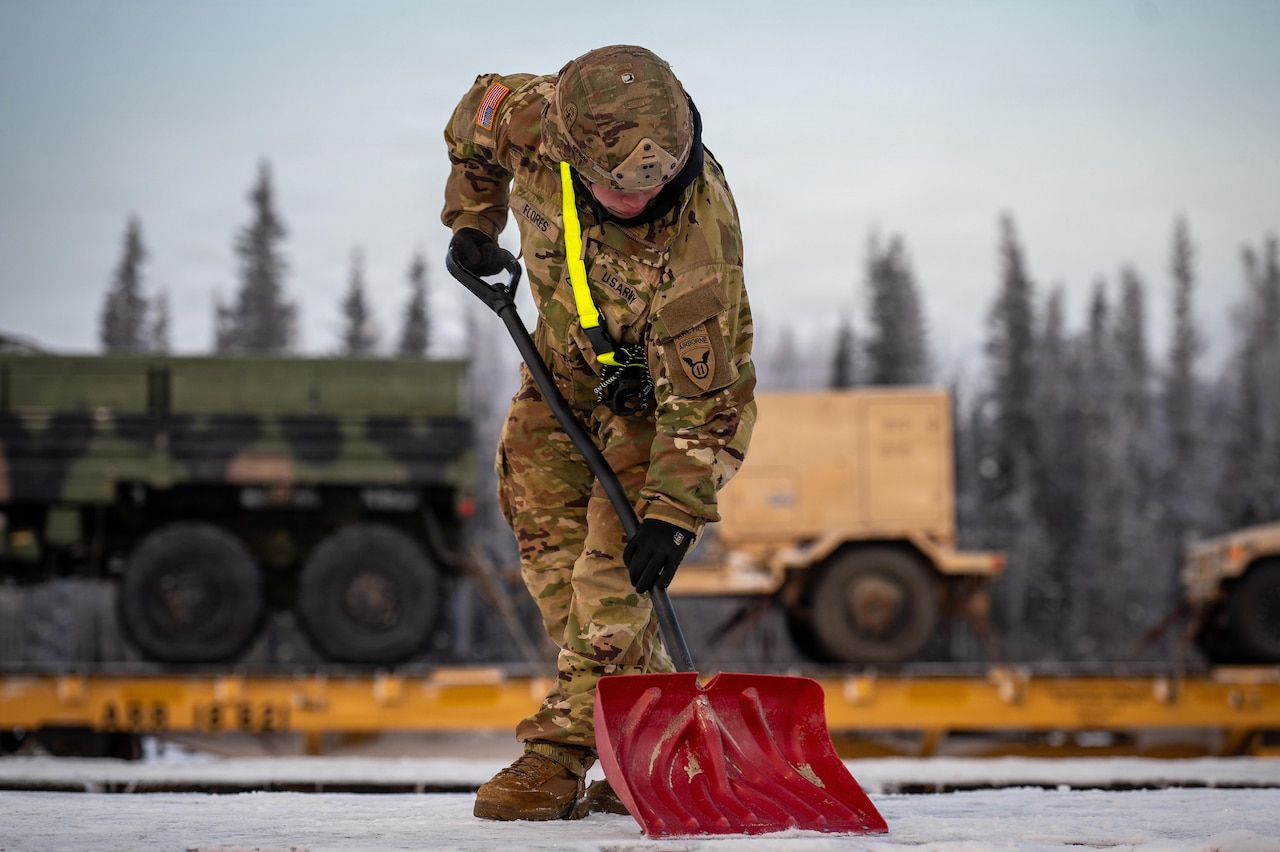 A man wearing a camouflage military uniform, safety belt and helmet shovels snow from the surface of a flatcar at a railhead; Another flatcar loaded with military vehicles is in the background.