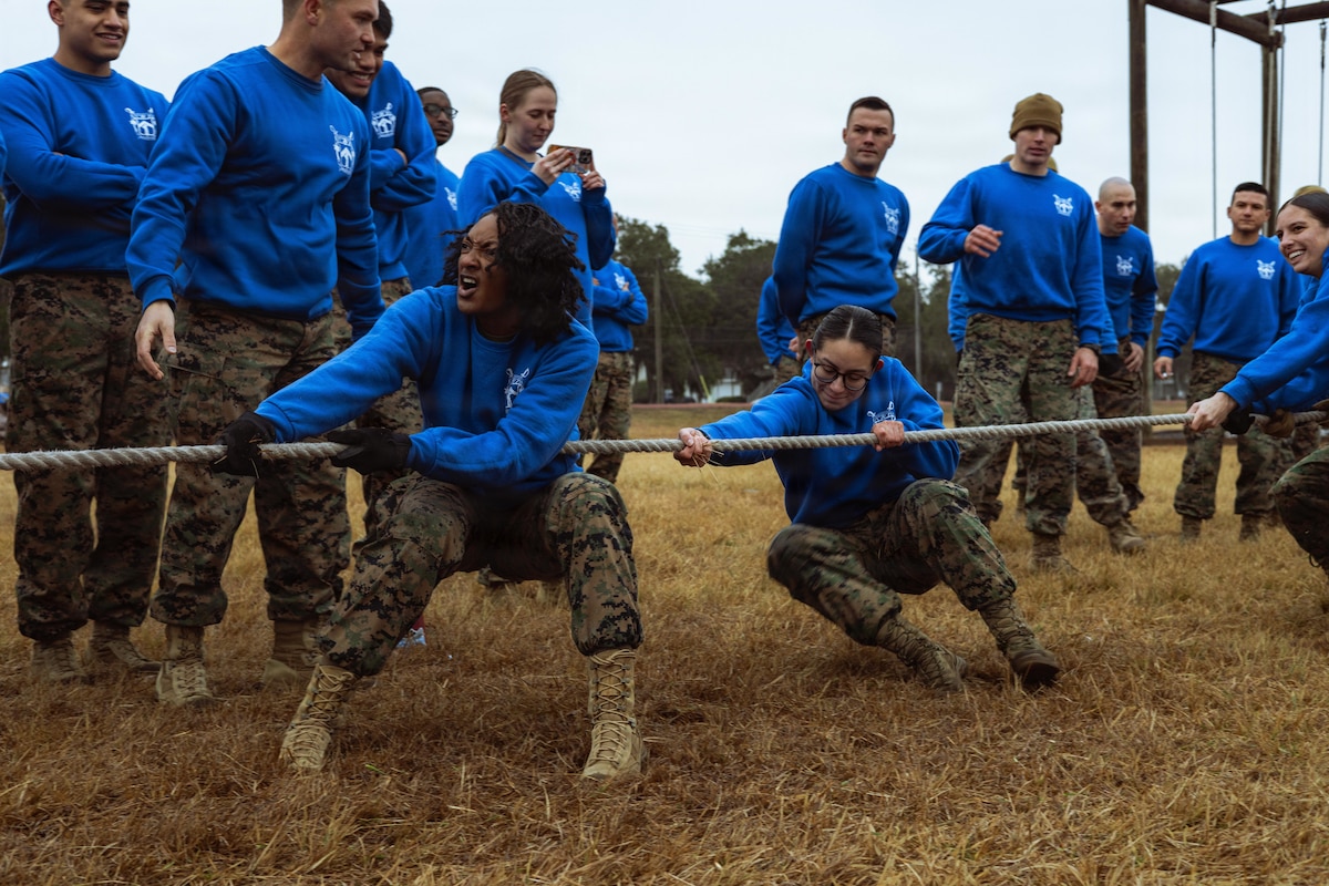 Three people wearing camouflaged pants and blue sweatshirts participate in a tug-of-war contest in a dry, grassy area as about a dozen people in similar attire observe.