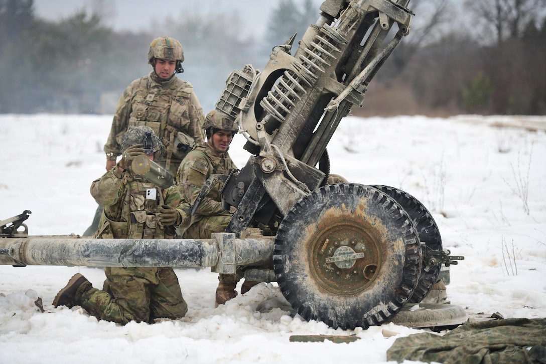 Three people wearing camouflage military uniforms, helmets and gloves kneel and stand in the snow next to a large military weapon, with trees in the background.