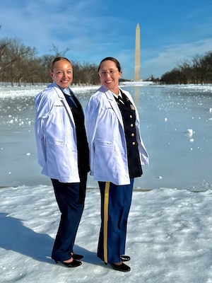 Two women service members wearing their white coats smile while standing next to each other.